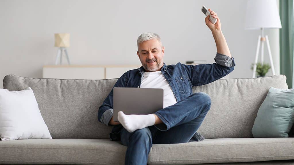 Happy senior man enjoying time at home, surfing on Internet, using notebook, holding remote for air conditioner