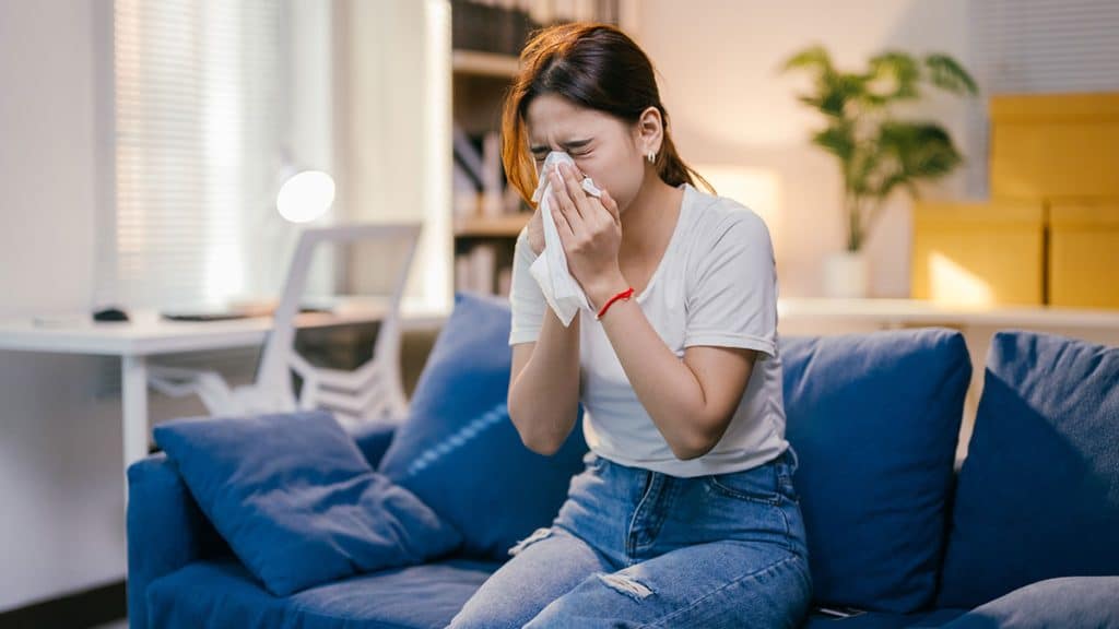 Young woman is blowing her nose with a tissue while sitting on a sofa in her living room